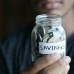A close-up image of a person's hand holding a jar full of coins labeled 'Savings'.
